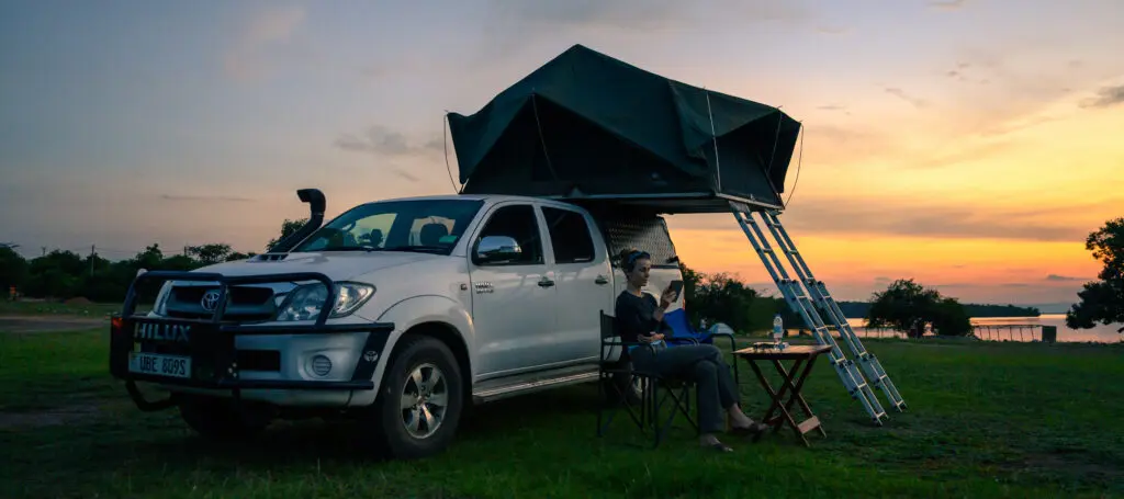 Young women reading in front of white Hilux with roof tent Lake Mburo campsite Uganda